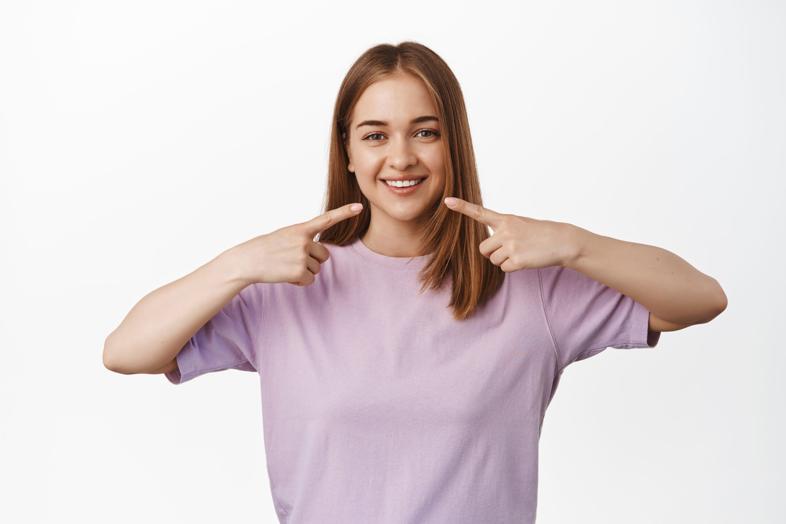Stomagology and dental clinic. Young blond woman in t-shirt pointing at her white teeth, mouth, standing against white background with big happy smile